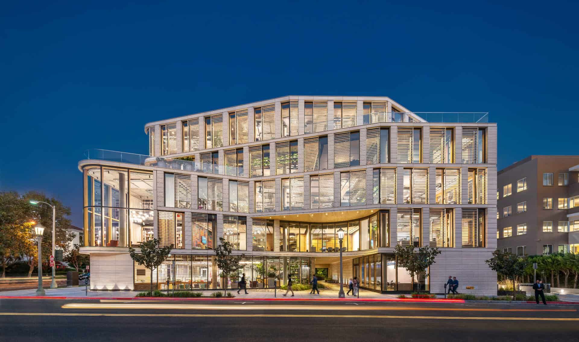Modern multi-story building with a light-colored, geometric grid facade, illuminated at twilight by a paved street.