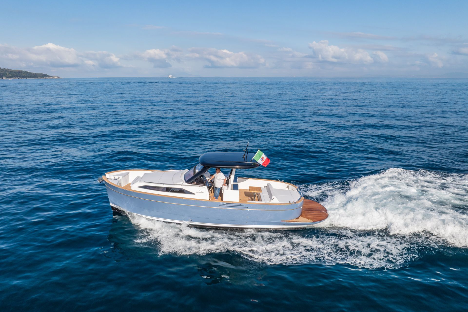 A light blue motorboat with a wooden deck cruises across calm blue water under a clear sky, displaying an Italian flag.