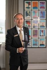 A professional sommelier in a suit holds a glass of red wine in front of a wall adorned with framed landscape prints.