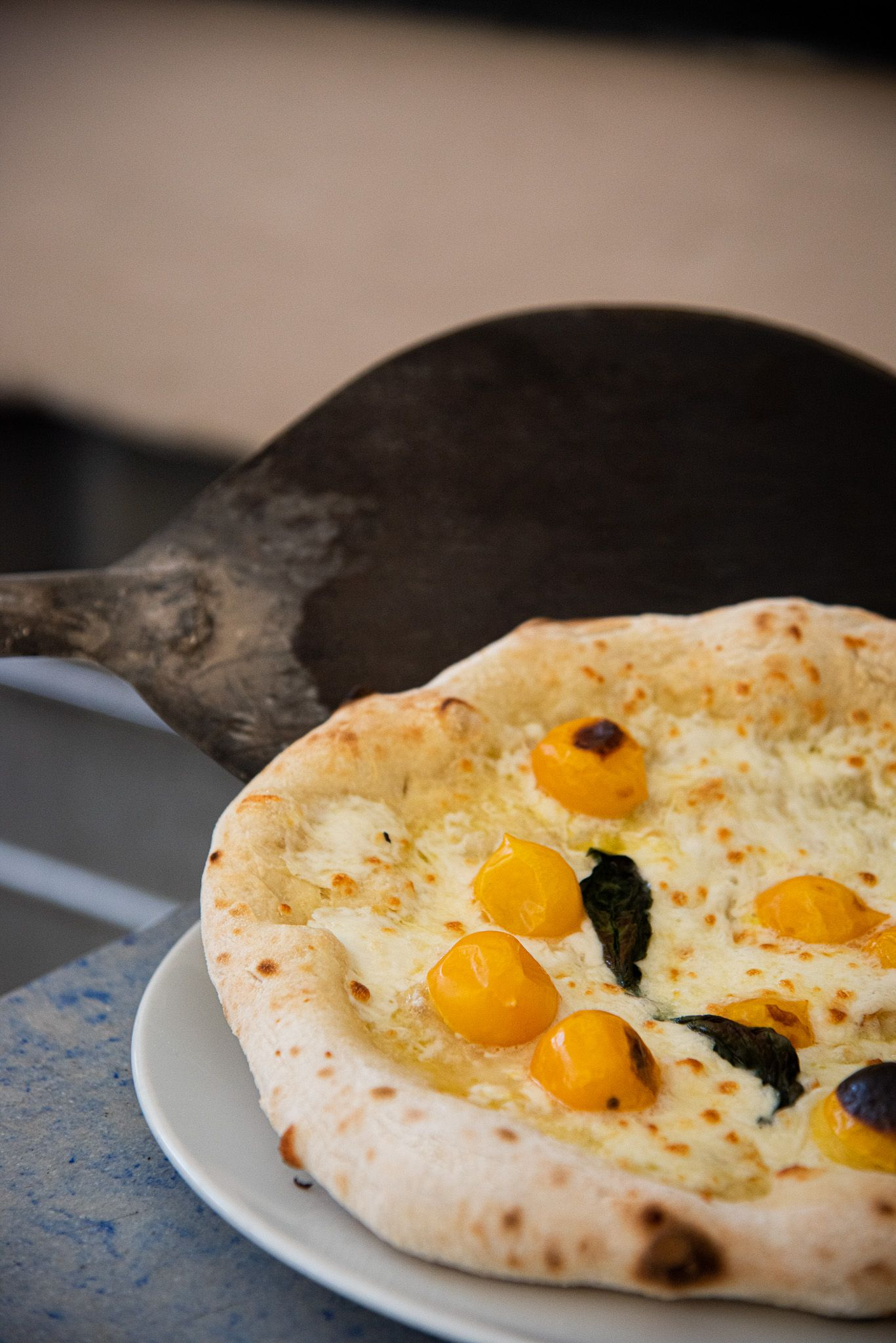 A wood-fired pizza with yellow cherry tomatoes and herbs on a white plate, held by a metal pizza peel.