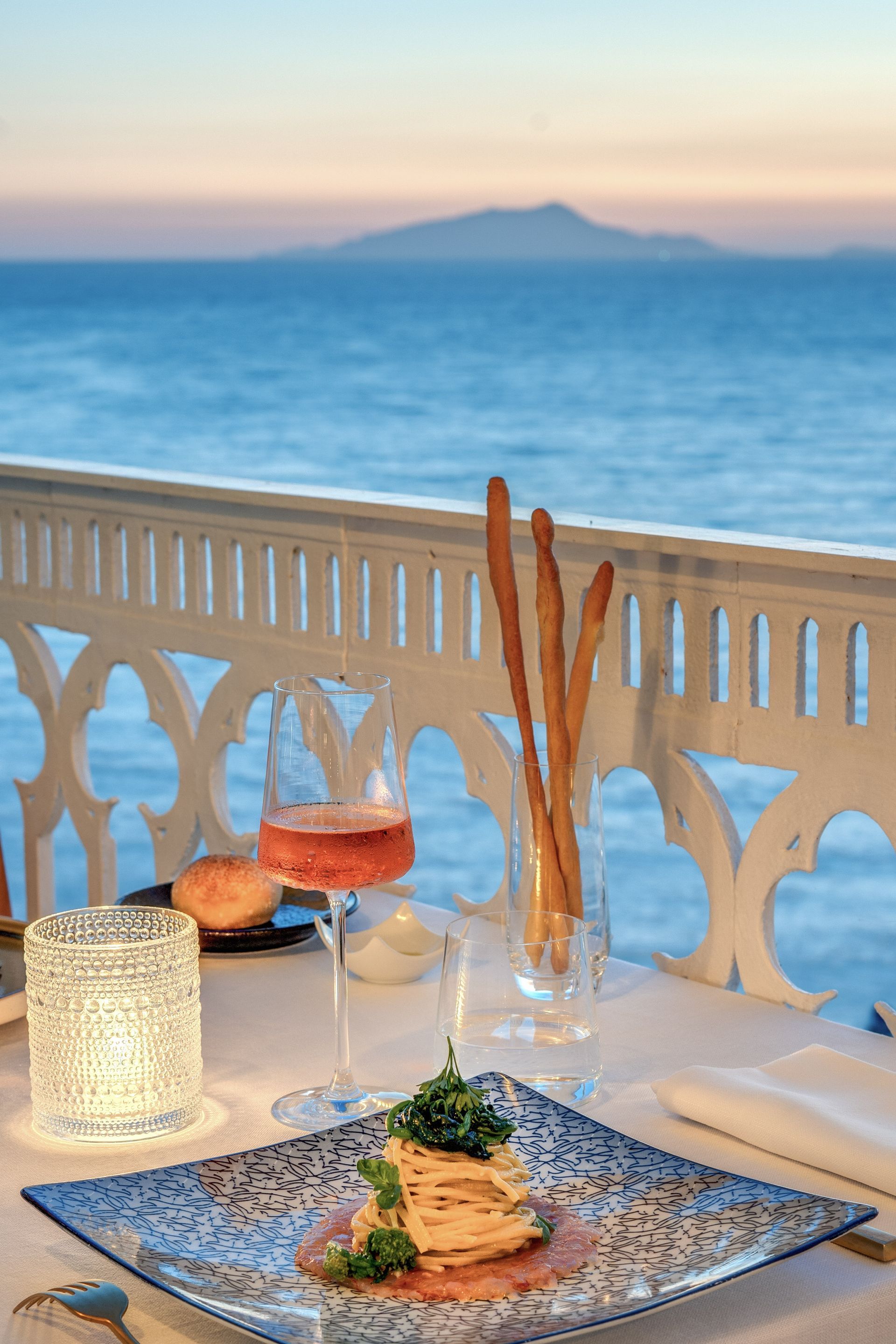 A meal with breadsticks and a glass of rosé on a table overlooking the sea at sunset with a distant island.