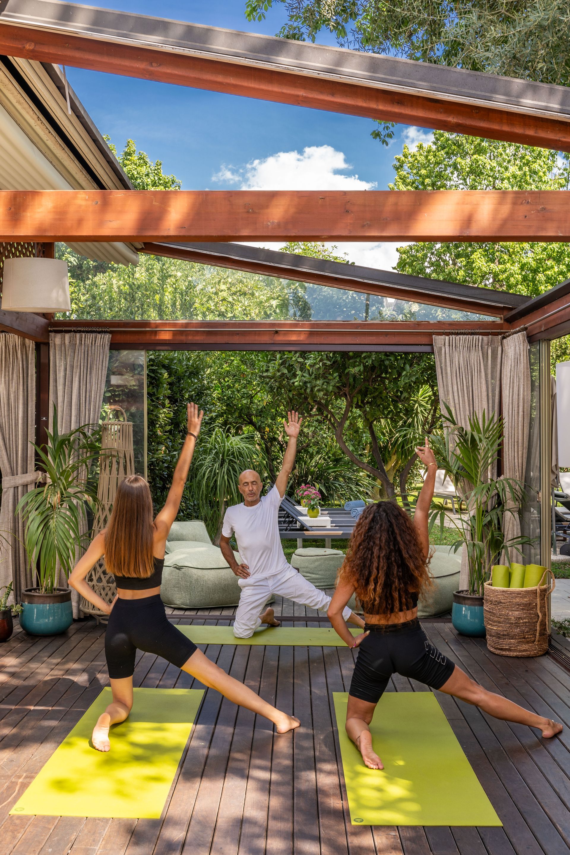 Three people in workout gear perform a kneeling side-stretch yoga pose on bright green mats in an open-air pavilion.