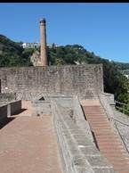A stone fortress wall with stairs leads toward a tall brick chimney set against a hillside under a clear blue sky.