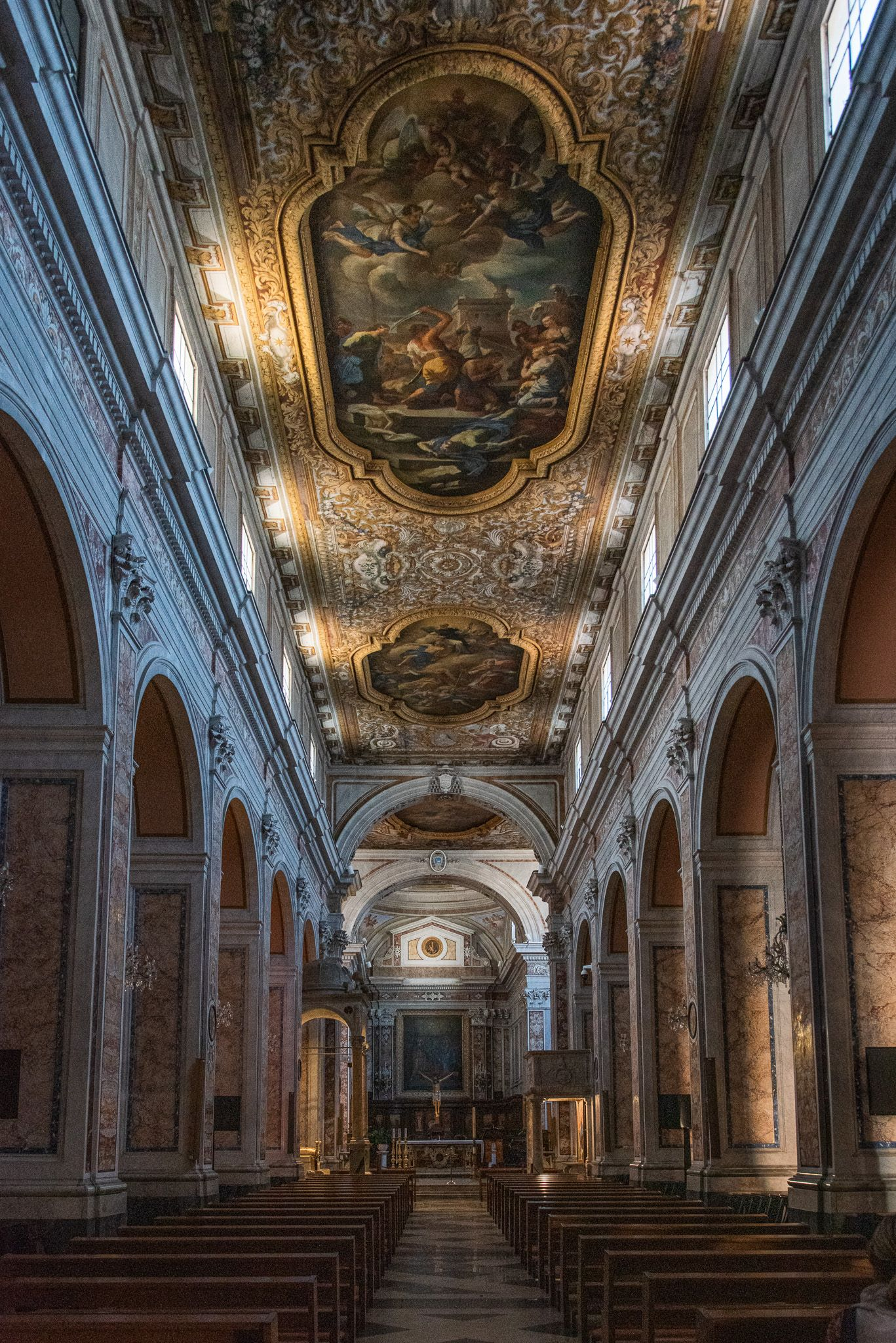 Ornate church interior with a long central aisle, arched pillars, and a ceiling decorated with large painted frescos.