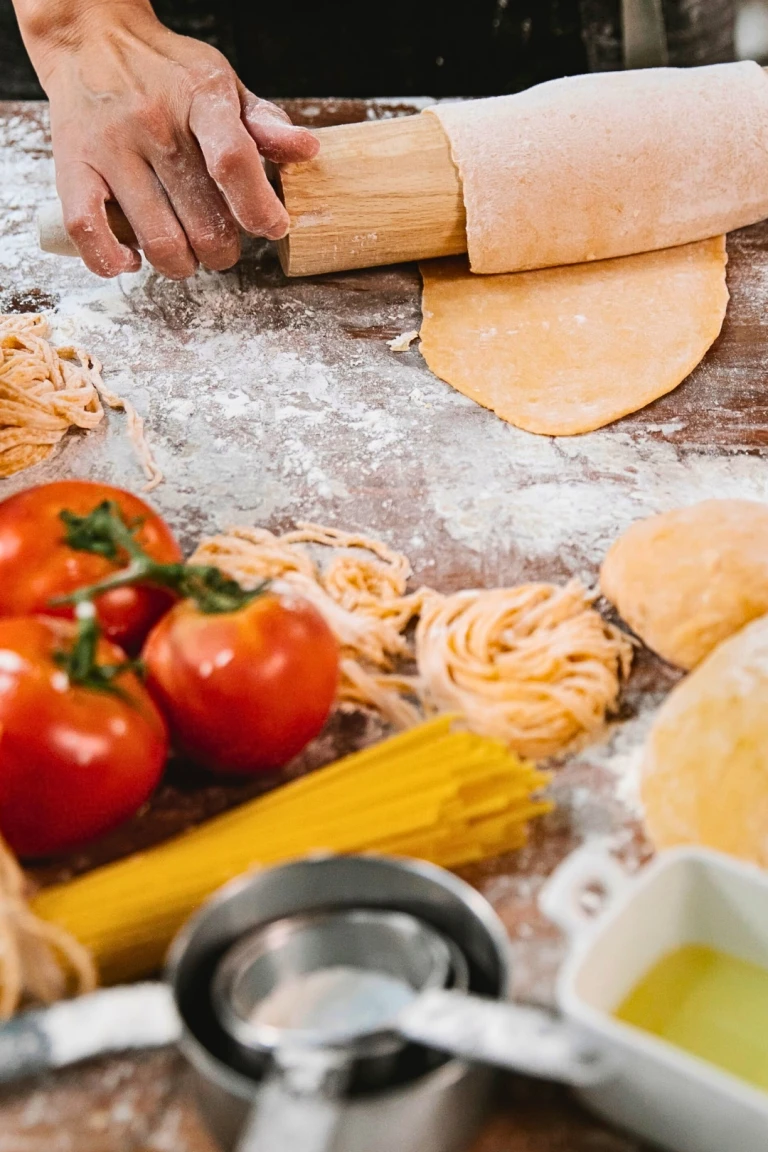 Hands roll fresh pasta dough on a flour-dusted counter near fresh tomatoes, dry pasta, and a bowl of oil.