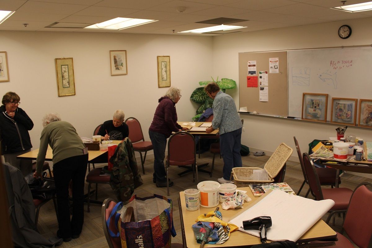 People crafting in a well-lit room, working at tables with supplies. Artwork displayed on walls and table.