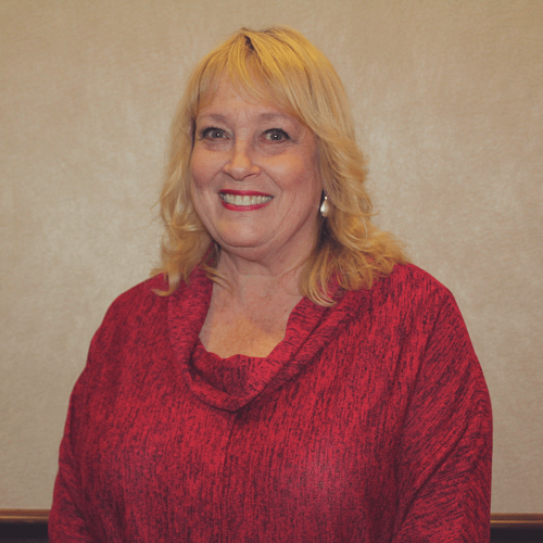 Woman with blonde hair wearing a red sweater smiles, standing in front of a neutral-colored wall.