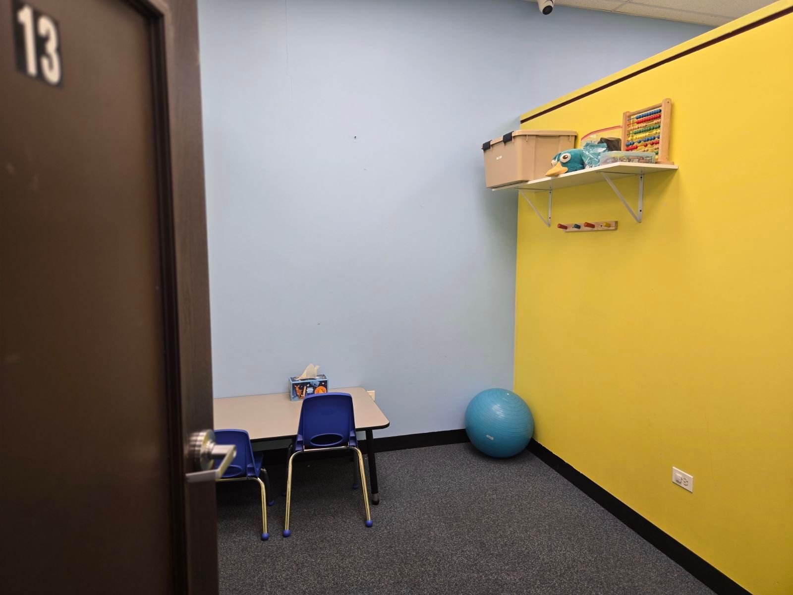 Small classroom corner with a table, chairs, shelves, and a blue exercise ball against yellow and gray walls.