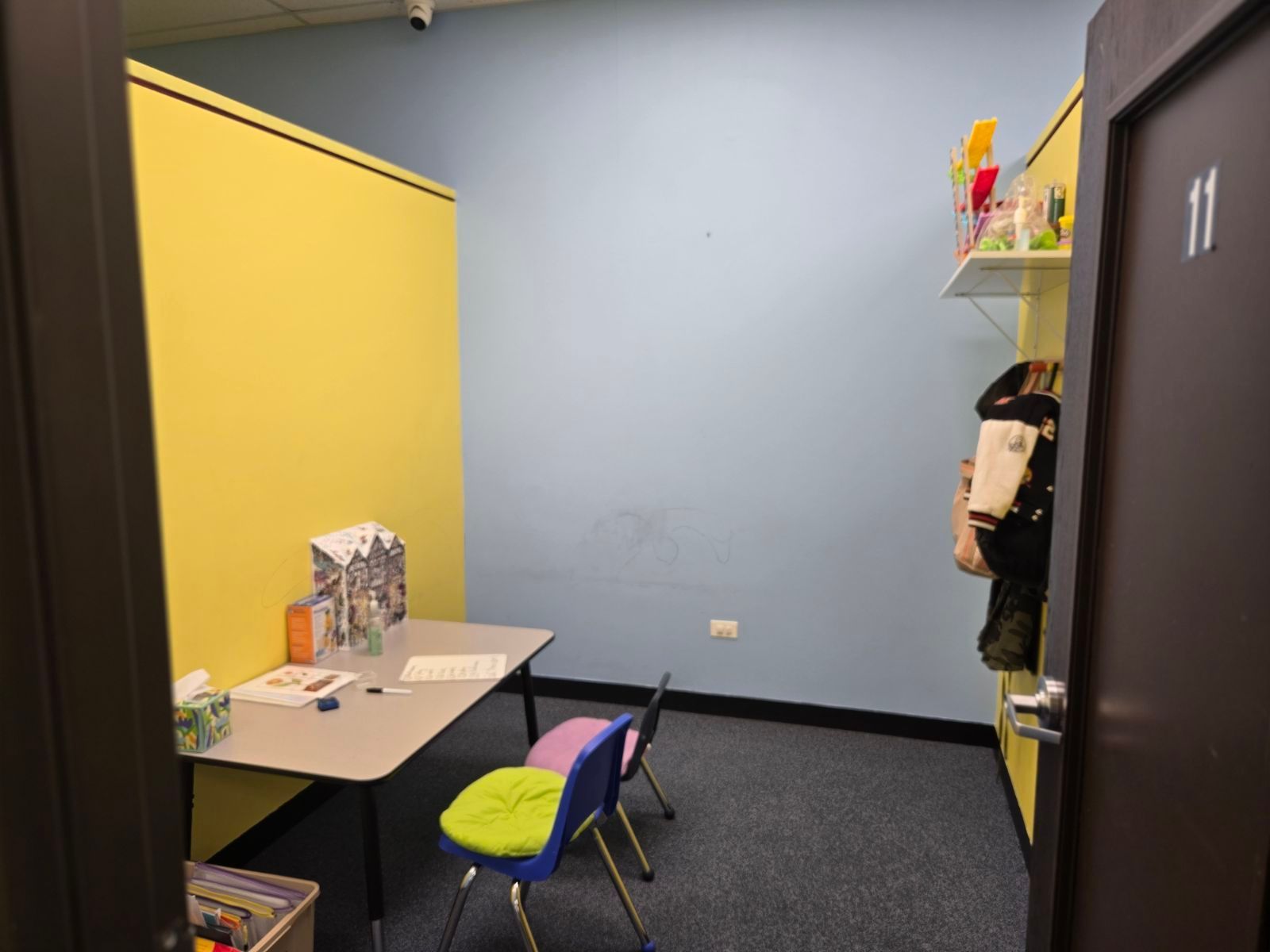 Small office nook with yellow wall, desk, chairs, shelves, and a person standing by the doorway.