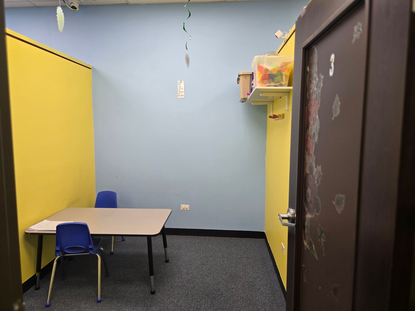 Small empty classroom with blue walls, a table and two blue chairs, and yellow cabinets/shelves.