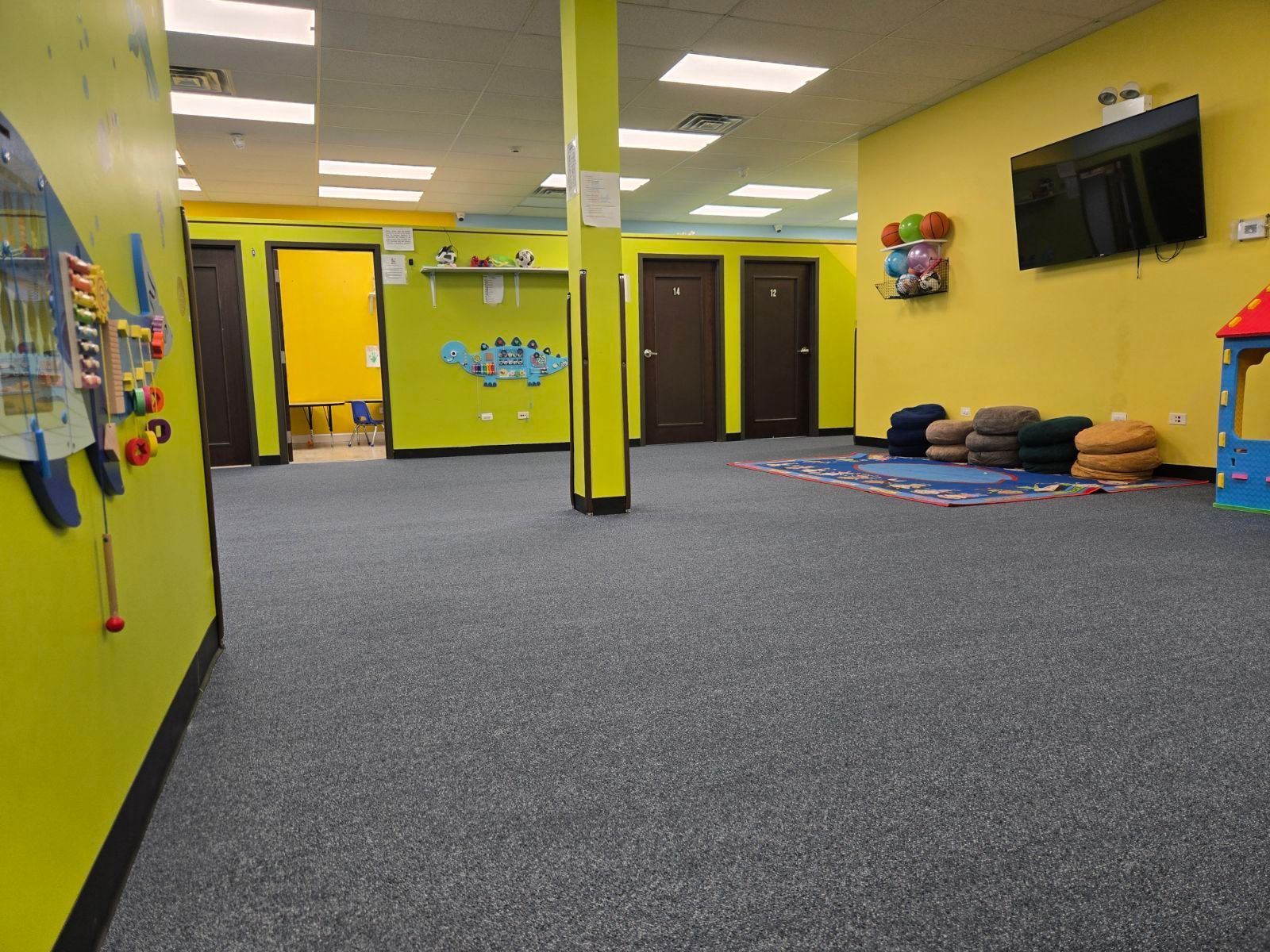 Bright yellow indoor room with carpet, columns, wall-mounted TV, and stacked mats near colorful doors.