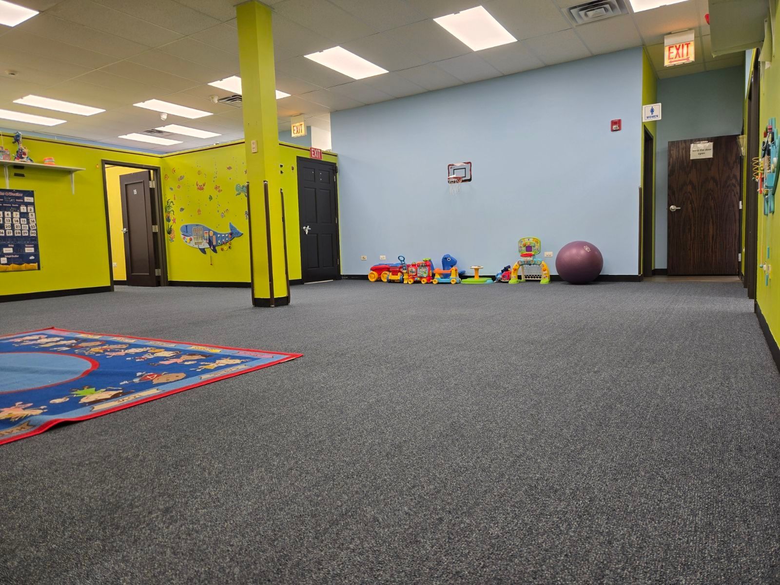 Empty daycare room with gray carpet, colorful wall murals, toys, and a purple exercise ball.