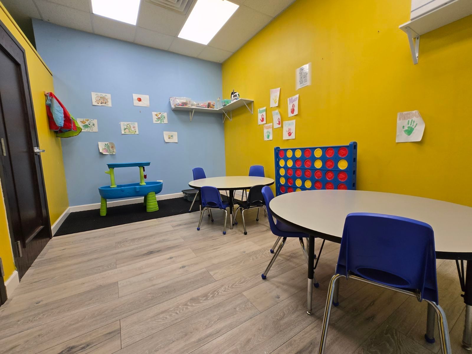 Colorful classroom with small tables, blue chairs, wall art, and a bright yellow and blue play area