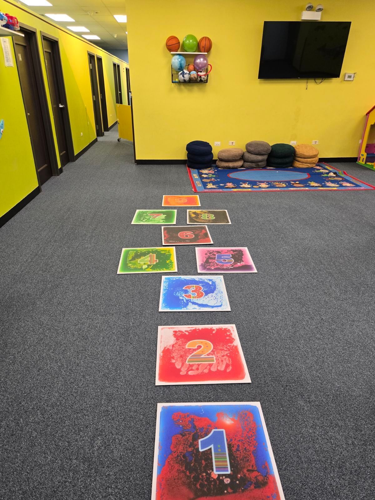 Colorful hopscotch-style floor path in a bright room with yellow walls, wall-mounted TV, and stacked cushions.