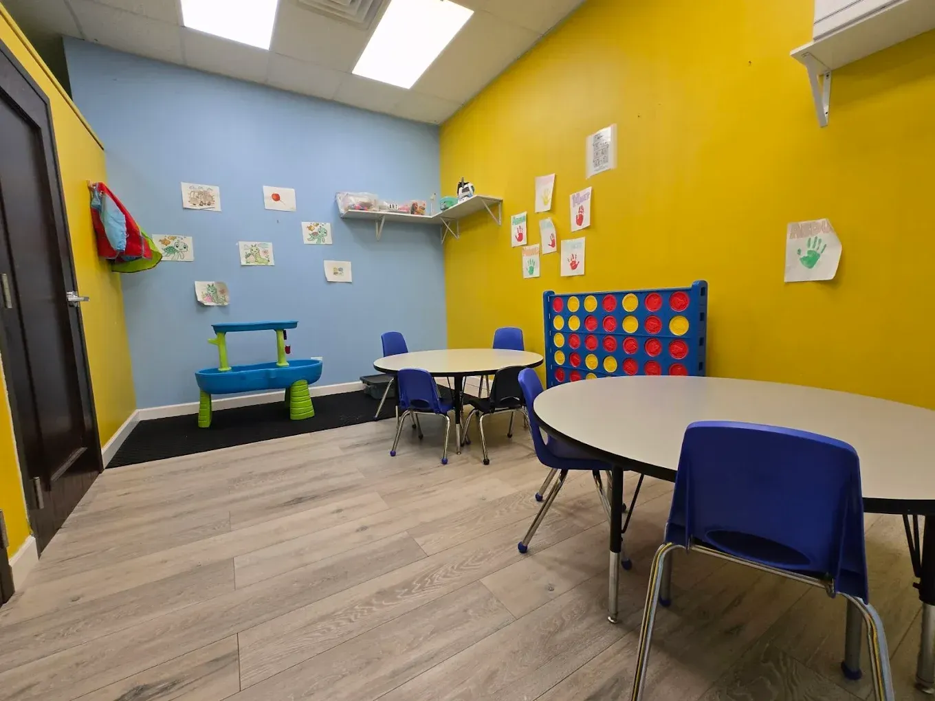 Colorful classroom with small tables, chairs, wall art, and a play area with toy shelves