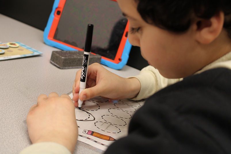 Child drawing with a marker on paper at a desk, with a tablet in a red-and-blue case nearby.