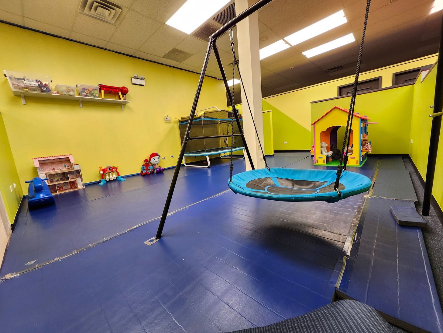 Indoor playroom with blue padded floor, trampoline, slide, and colorful toys against yellow walls