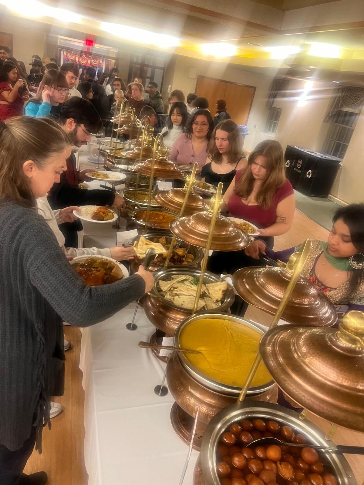 People serving food at a long buffet table in a bright dining hall with copper pots and bowls
