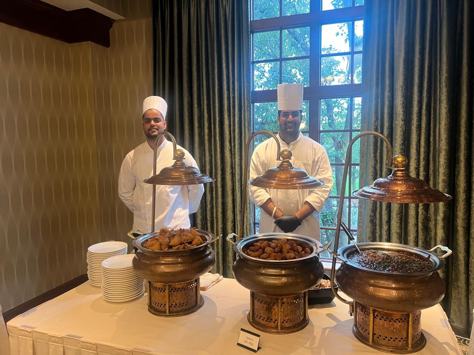 Two chefs in white uniforms stand behind a buffet with covered chafing dishes and plates.
