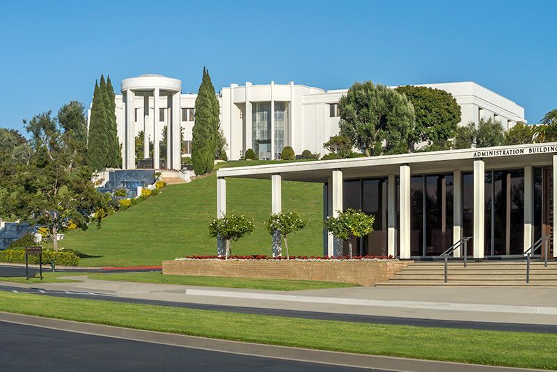 White building complex on a grassy hill with trees; a sunny day.