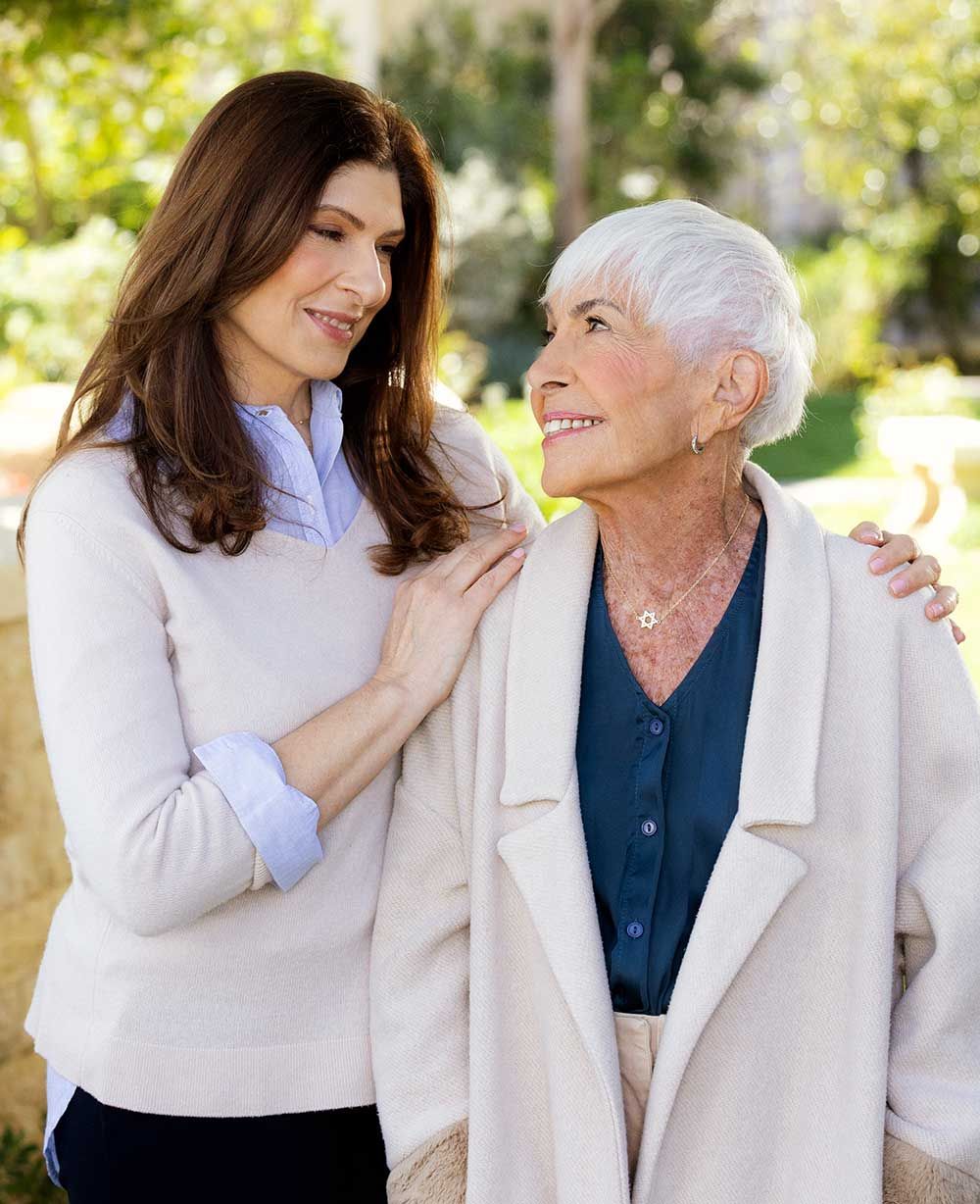 Two women, one younger and one older, smiling at each other in a garden.