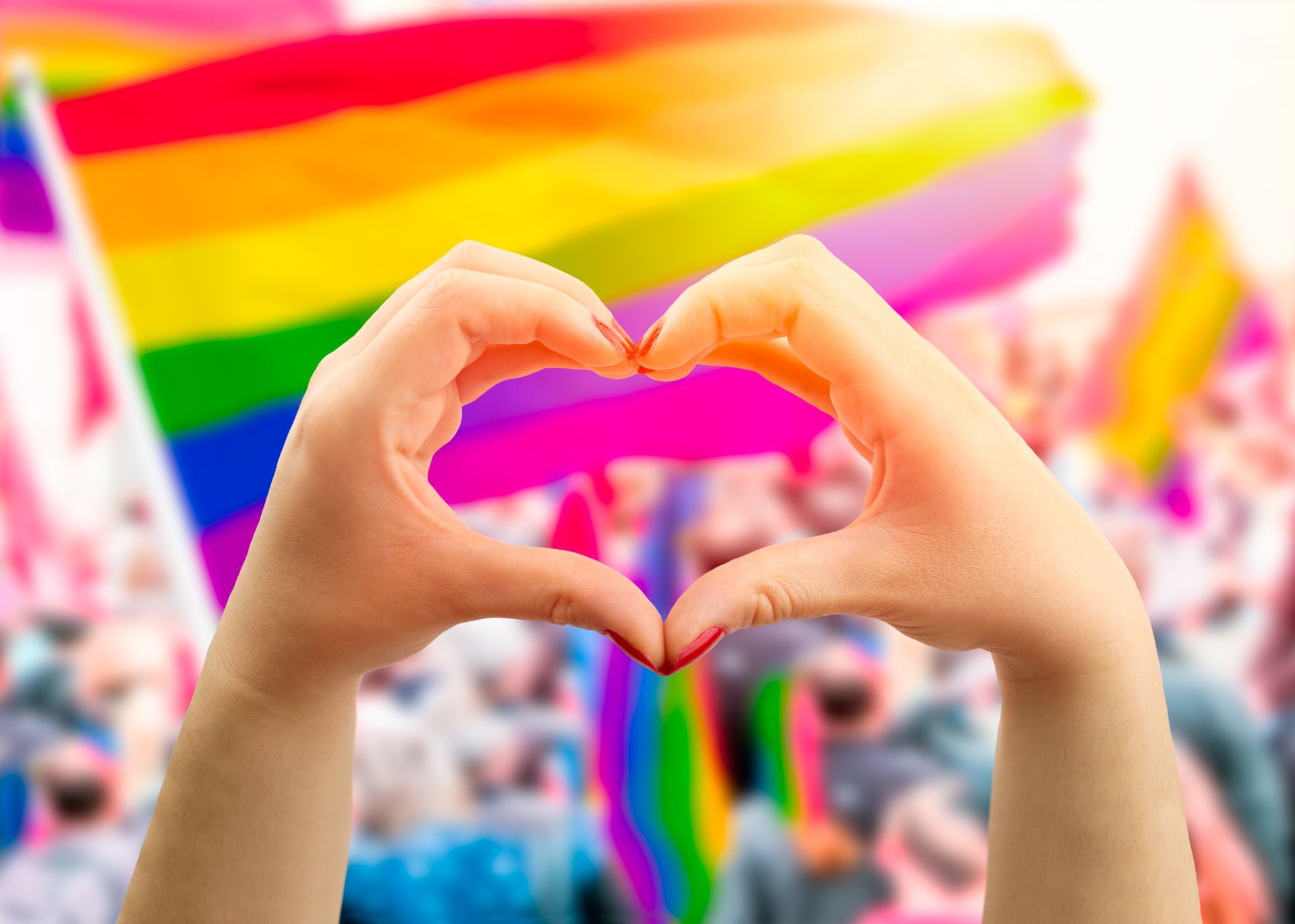 A person is making a heart shape with their hands in front of a rainbow flag.