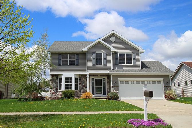 Two-story gray suburban house with a front lawn, driveway, and white garage under a blue sky.