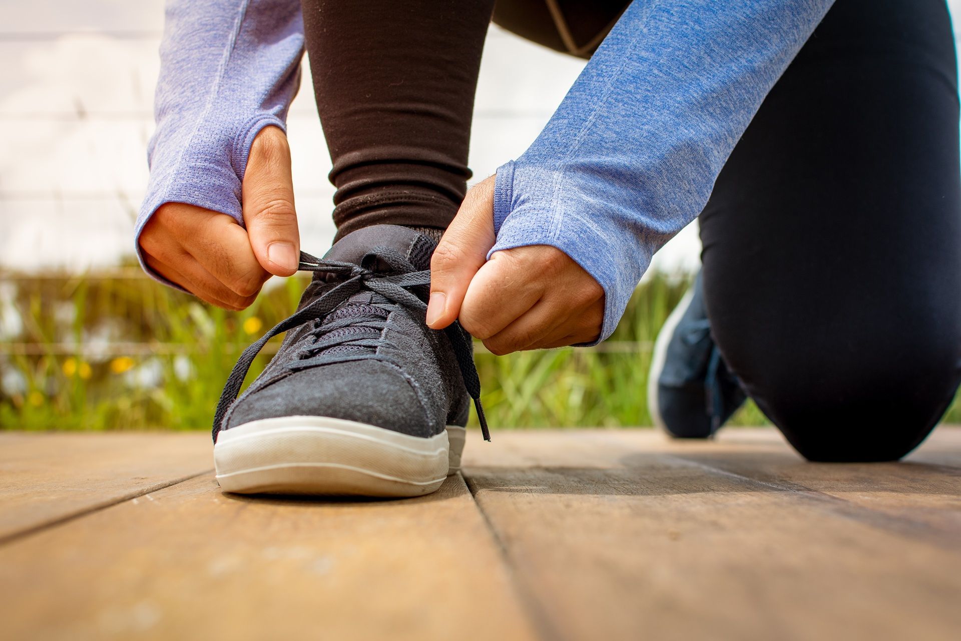 Person kneeling, tying shoelace of a gray sneaker on a wooden deck, wearing blue workout gear.