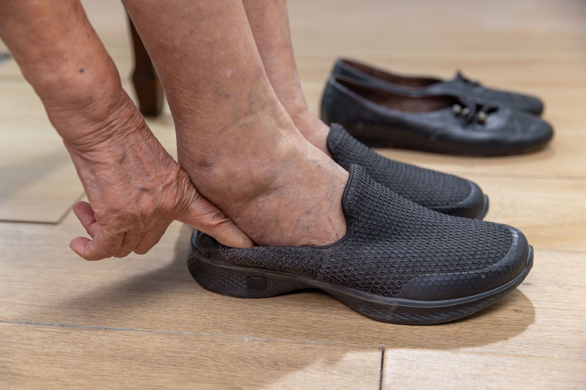 Person putting on a black slip-on shoe. Showing swollen ankles. Neutral background.