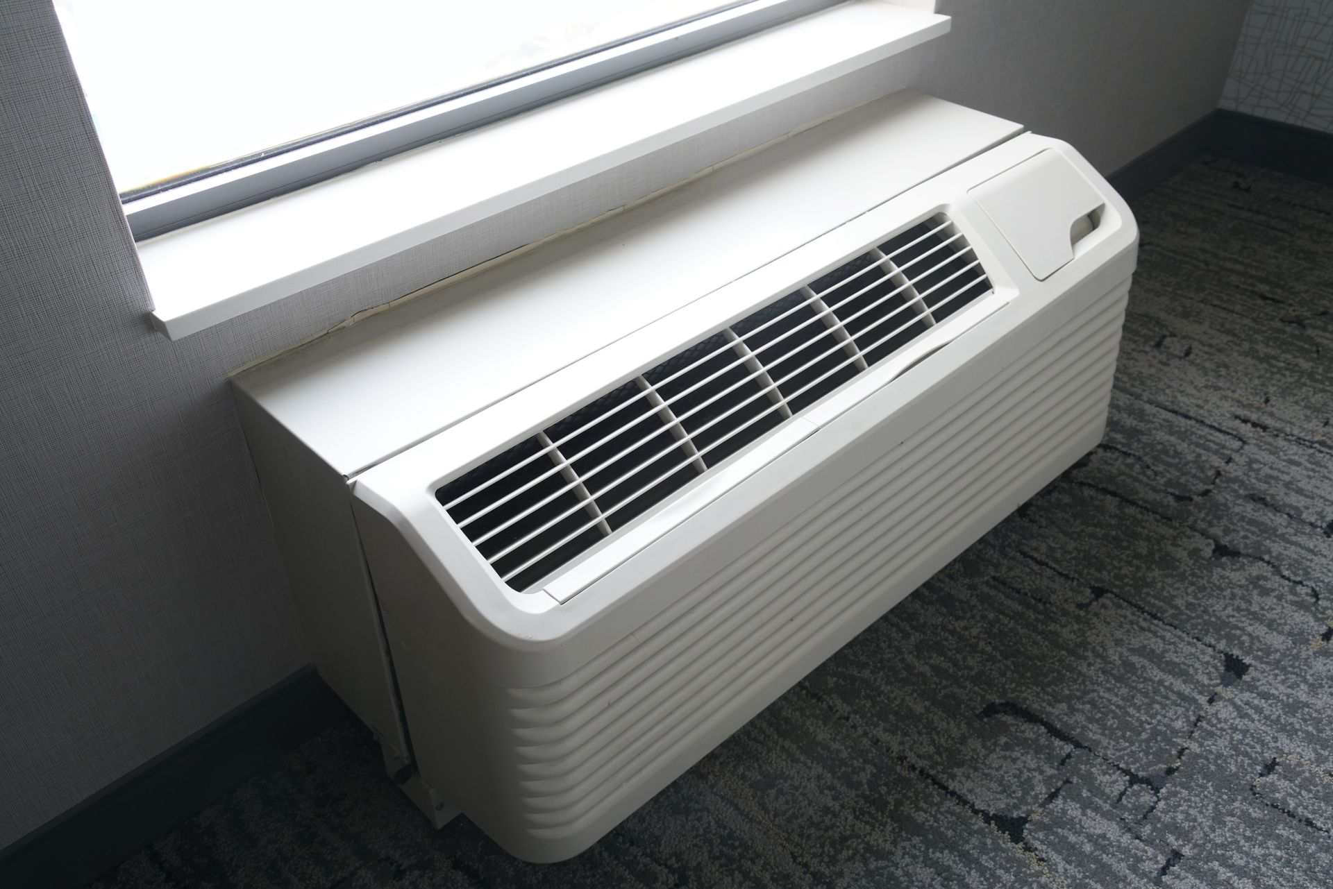 A white PTAC HVAC unit is installed under a window in a room with gray carpet and textured walls.