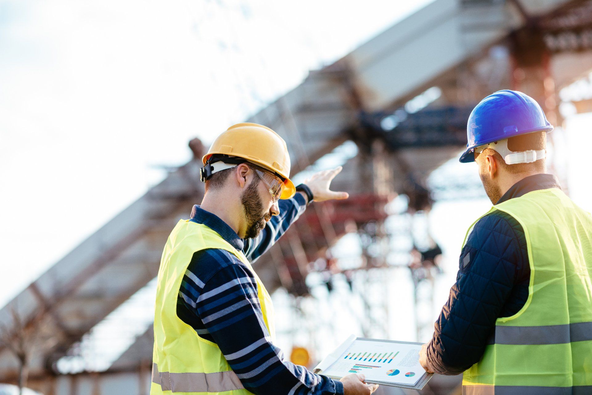 Two construction workers in safety vests and hard hats reviewing a document at a construction site.
