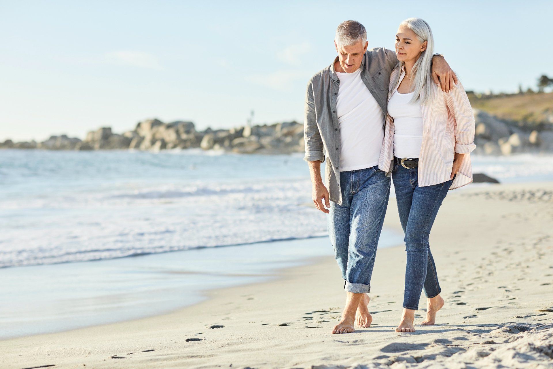 Couple walks barefoot on a beach, man's arm around woman. Ocean and coastline in background.