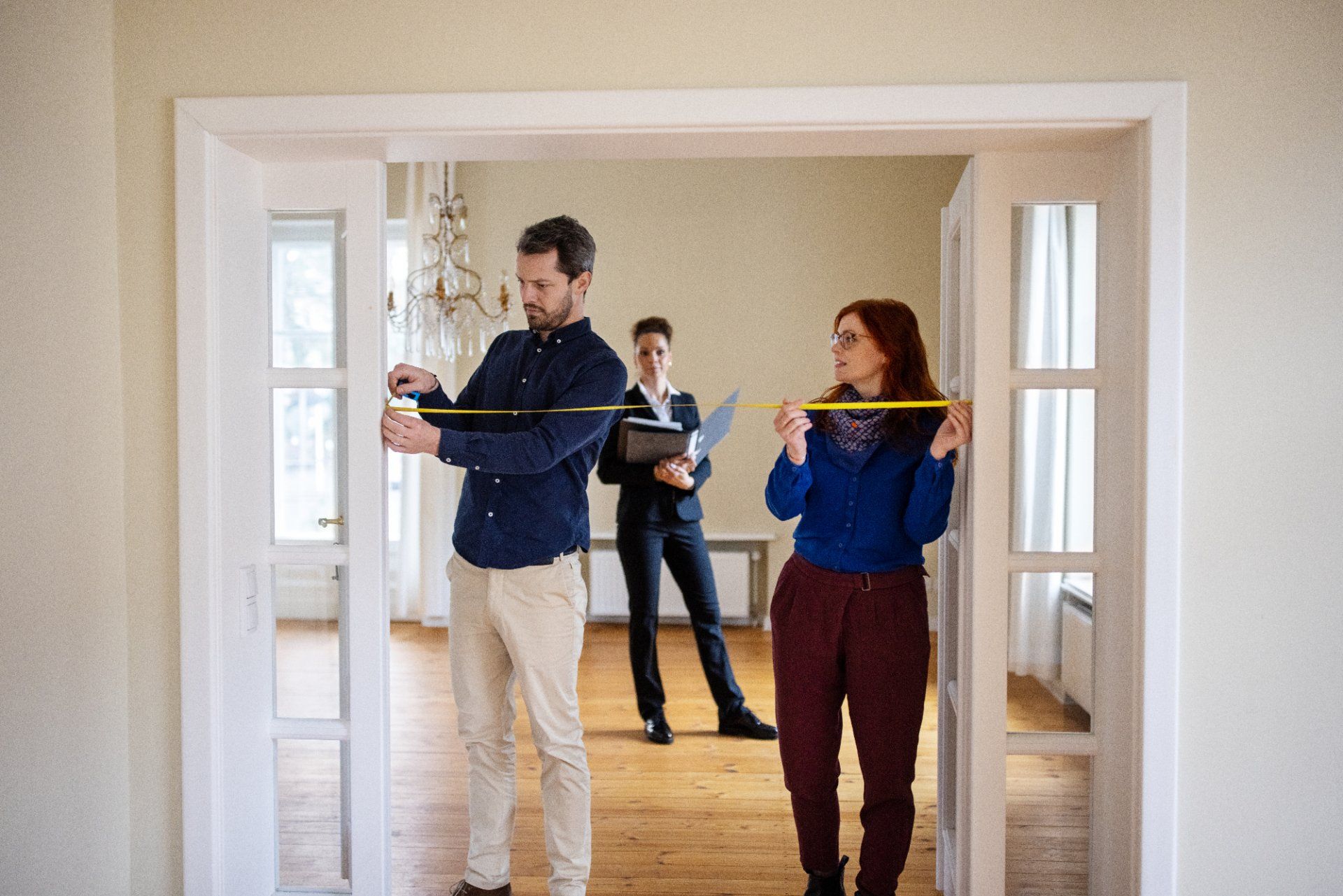 Two people measure a doorway; a third person in a suit observes. They are indoors in a room with hardwood floors.