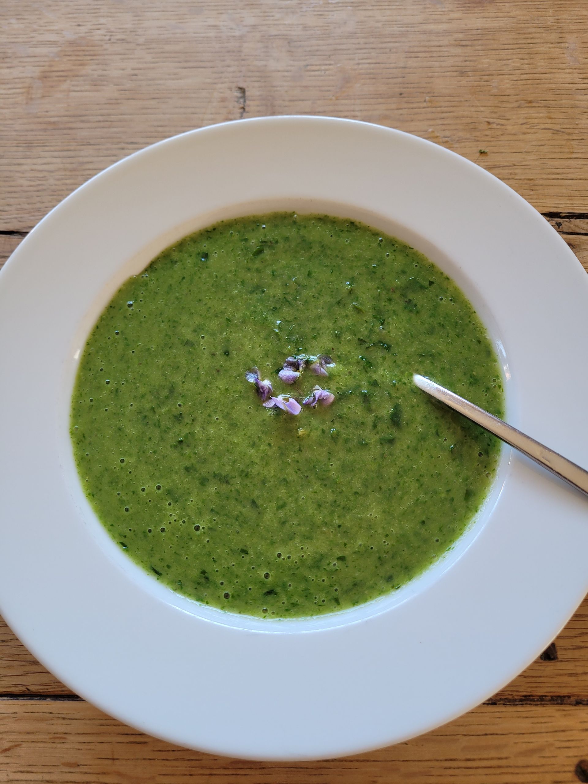 Groene soep in een witte kom met een lepel, gegarneerd met kleine paarse bloemen, op een houten tafel.