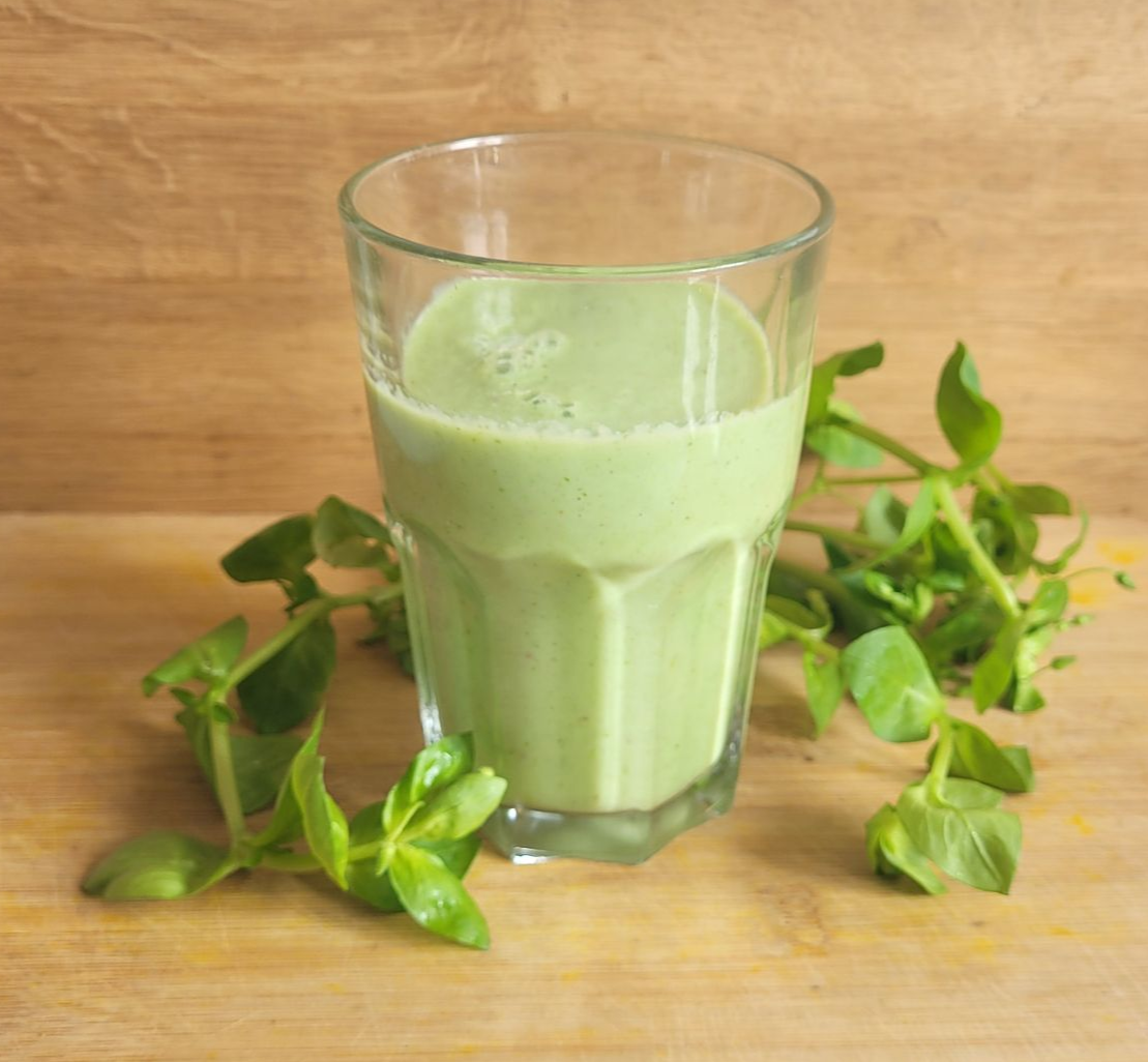 Een groene smoothie in een glas op een houten ondergrond, met bladgroenten ernaast.