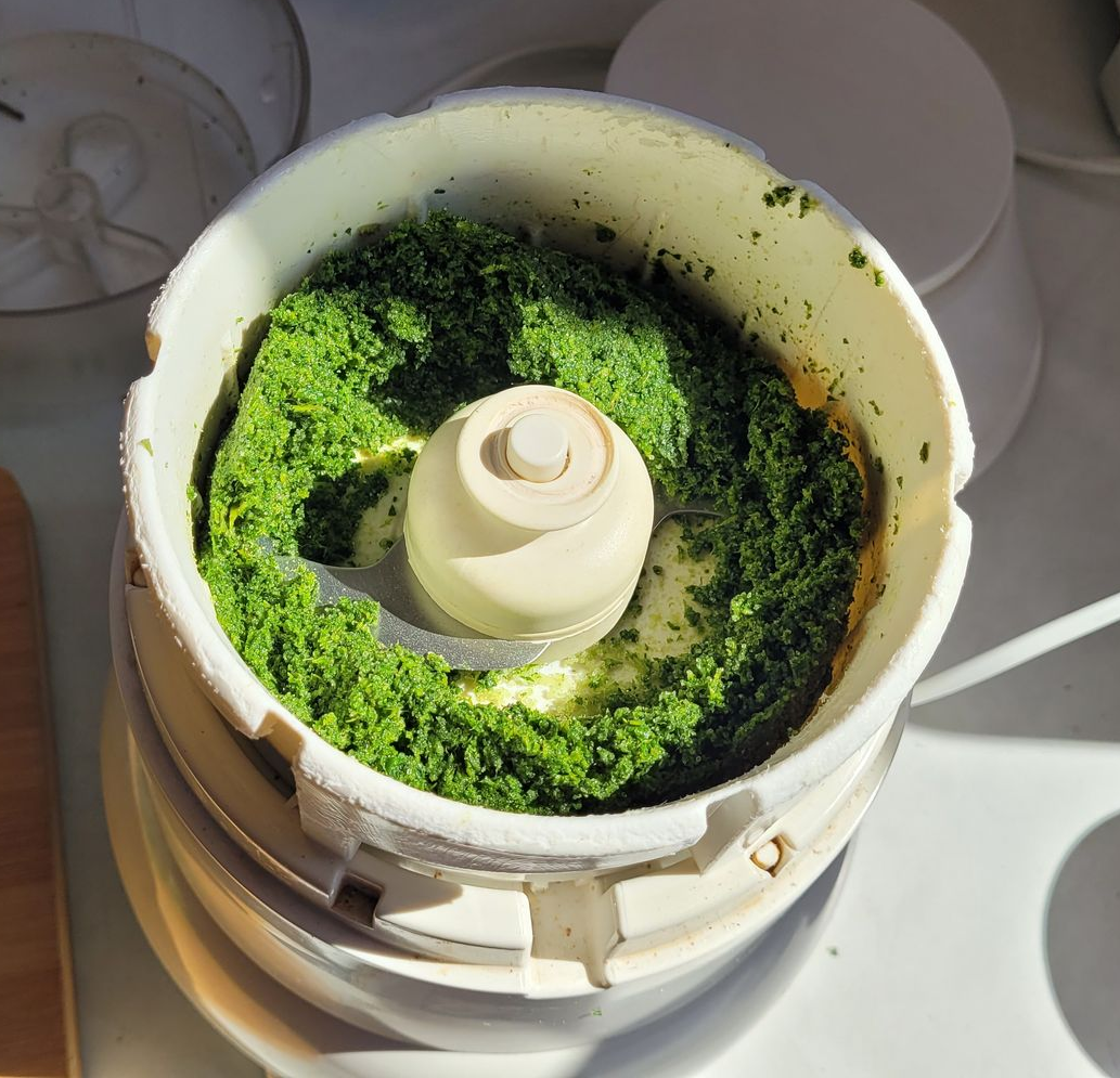 Bovenaanzicht van een witte blenderkom met groene, fijngehakte kruiden of puree rond de centrale messenas.
