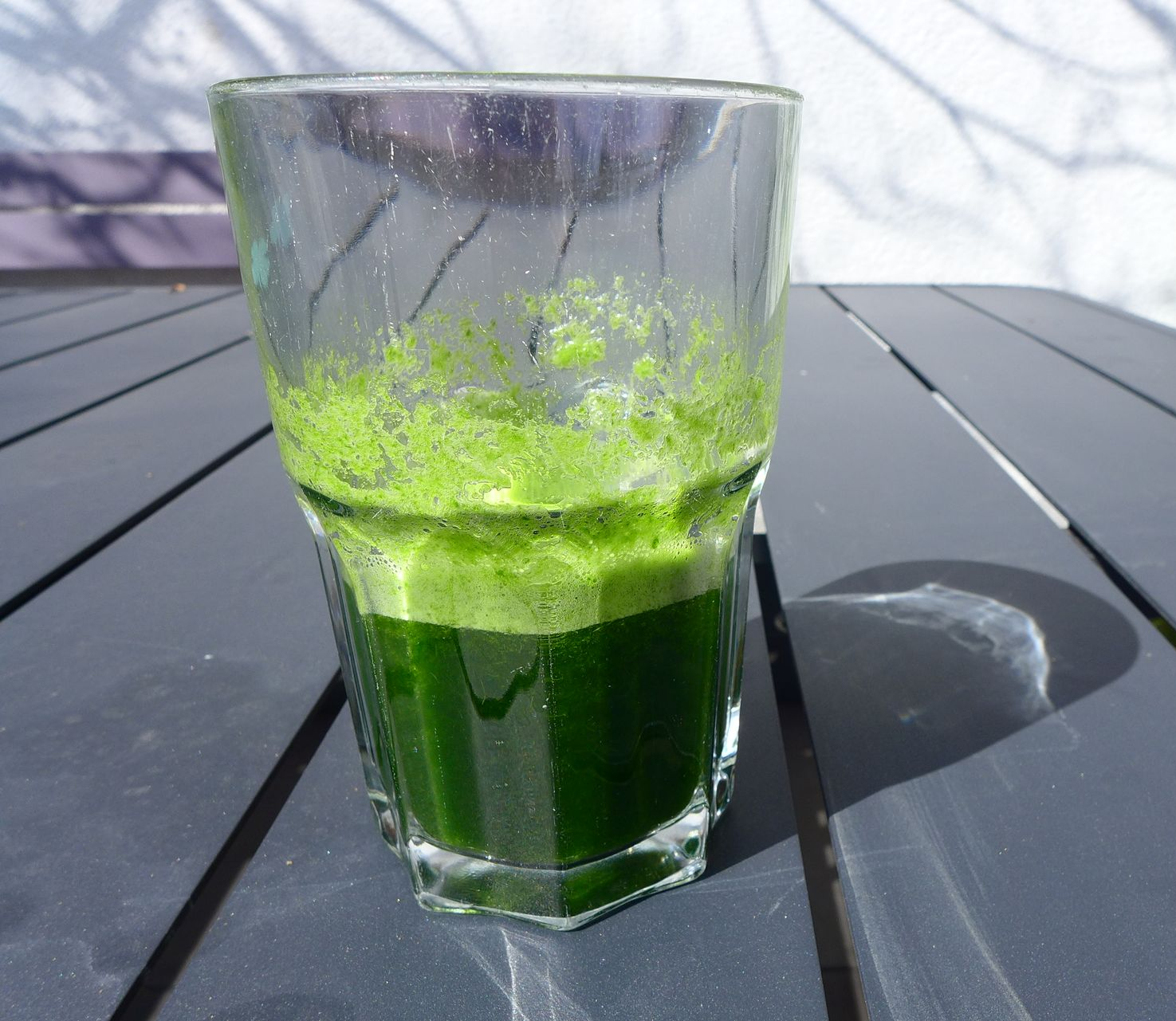Een glas met felgroene vloeistof en schuim staat op een donkere terrastafel in de zon.
