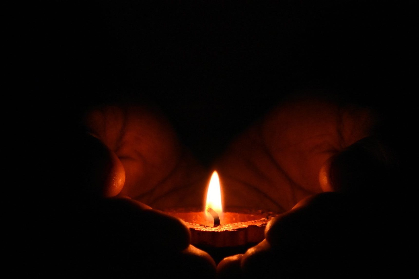 Handen omsluiten een brandende kaars in het donker, met een warme oranje vlam die gloeit.