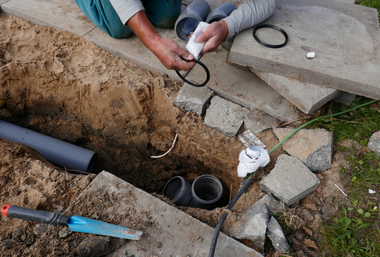 Person installing plumbing in a trench, holding an O-ring and pipe, surrounded by dirt, concrete slabs, and tools.