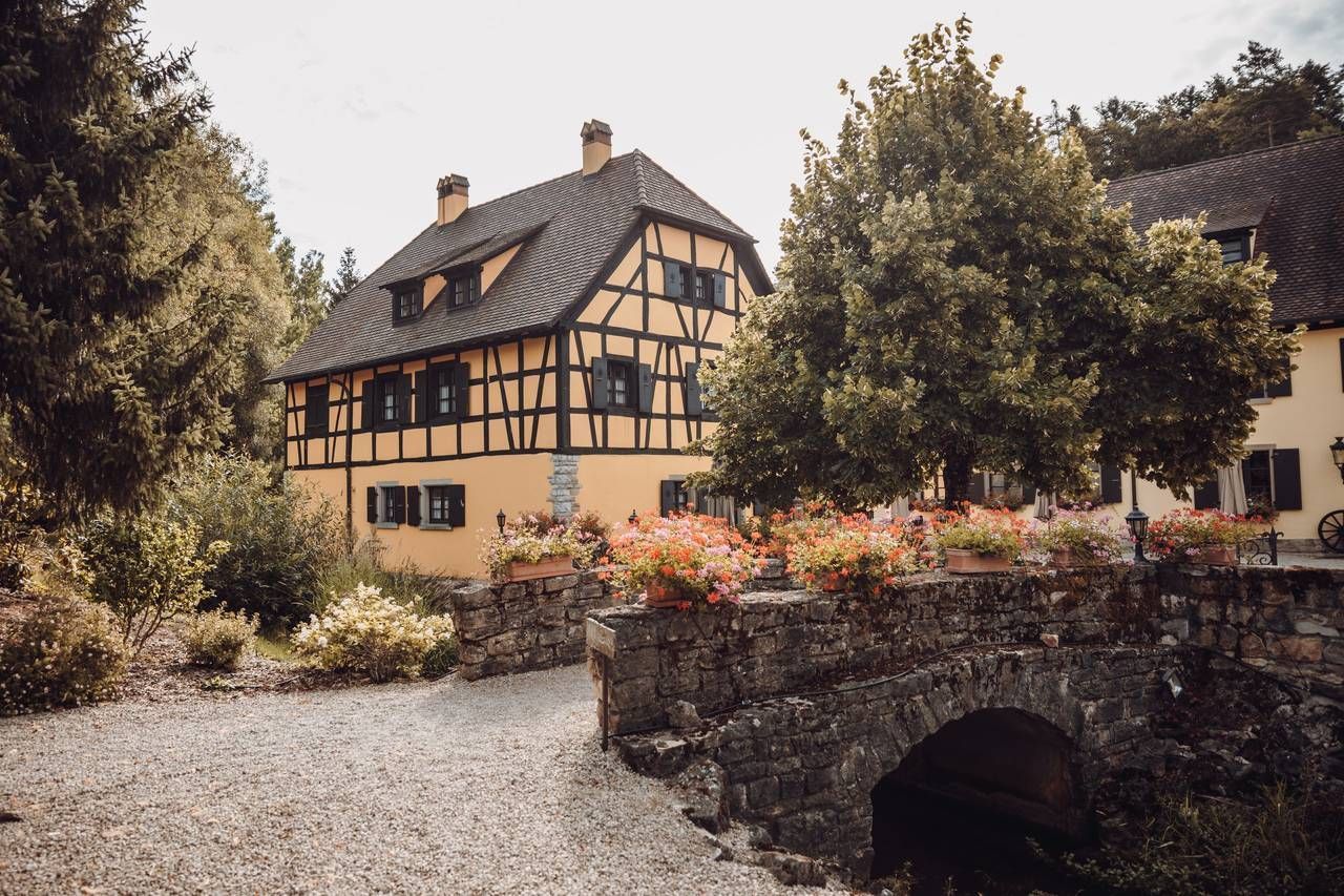 Une maison jaune avec un toit noir est entourée d'arbres et d'un pont de pierre.
