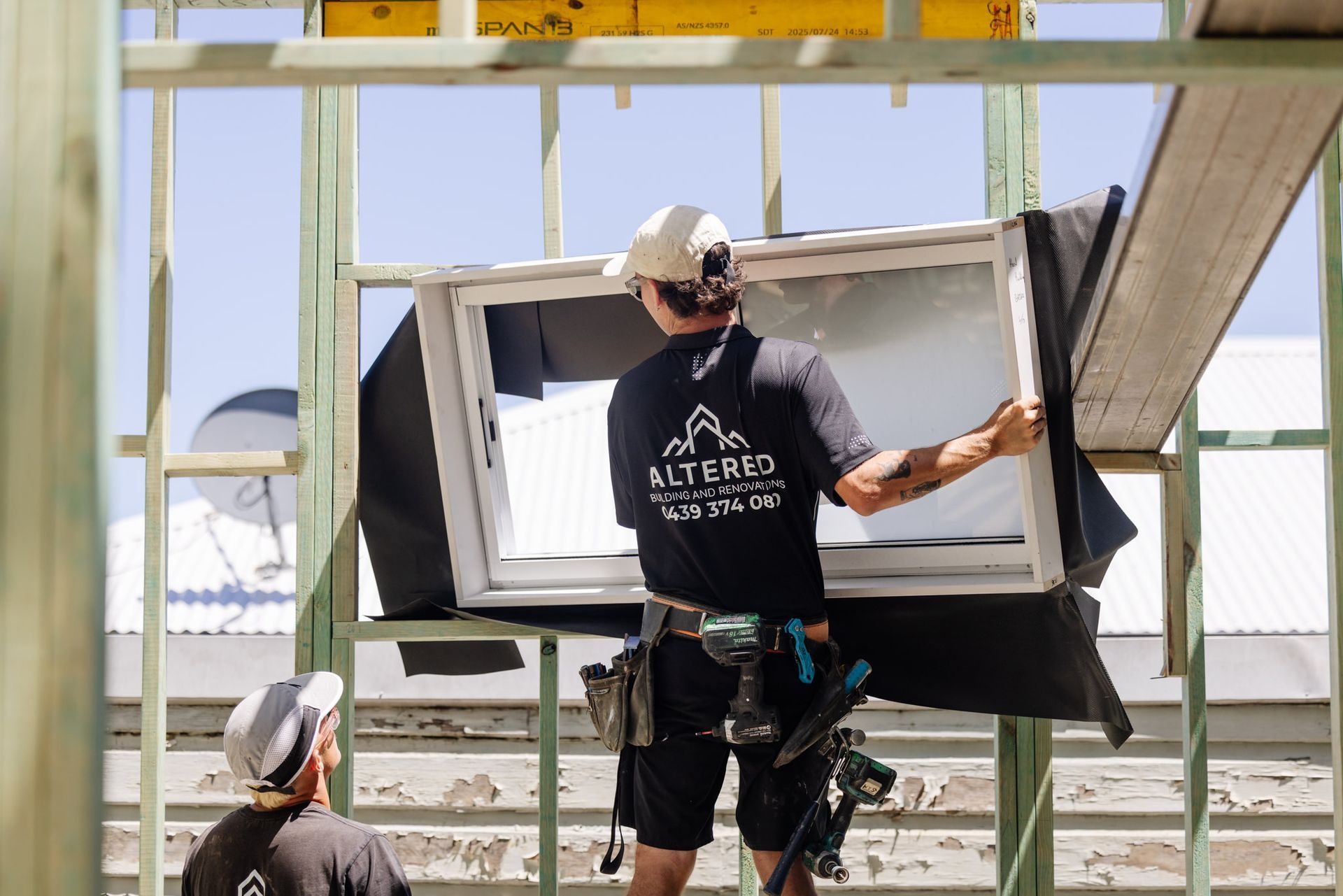 Two construction workers in black uniforms install a white window frame into a wooden building frame.