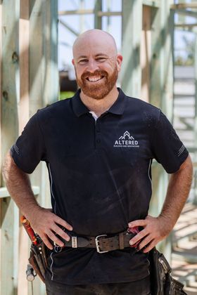 A smiling person with a red beard wearing a black polo shirt and tool belt, standing in a partially built wooden frame.