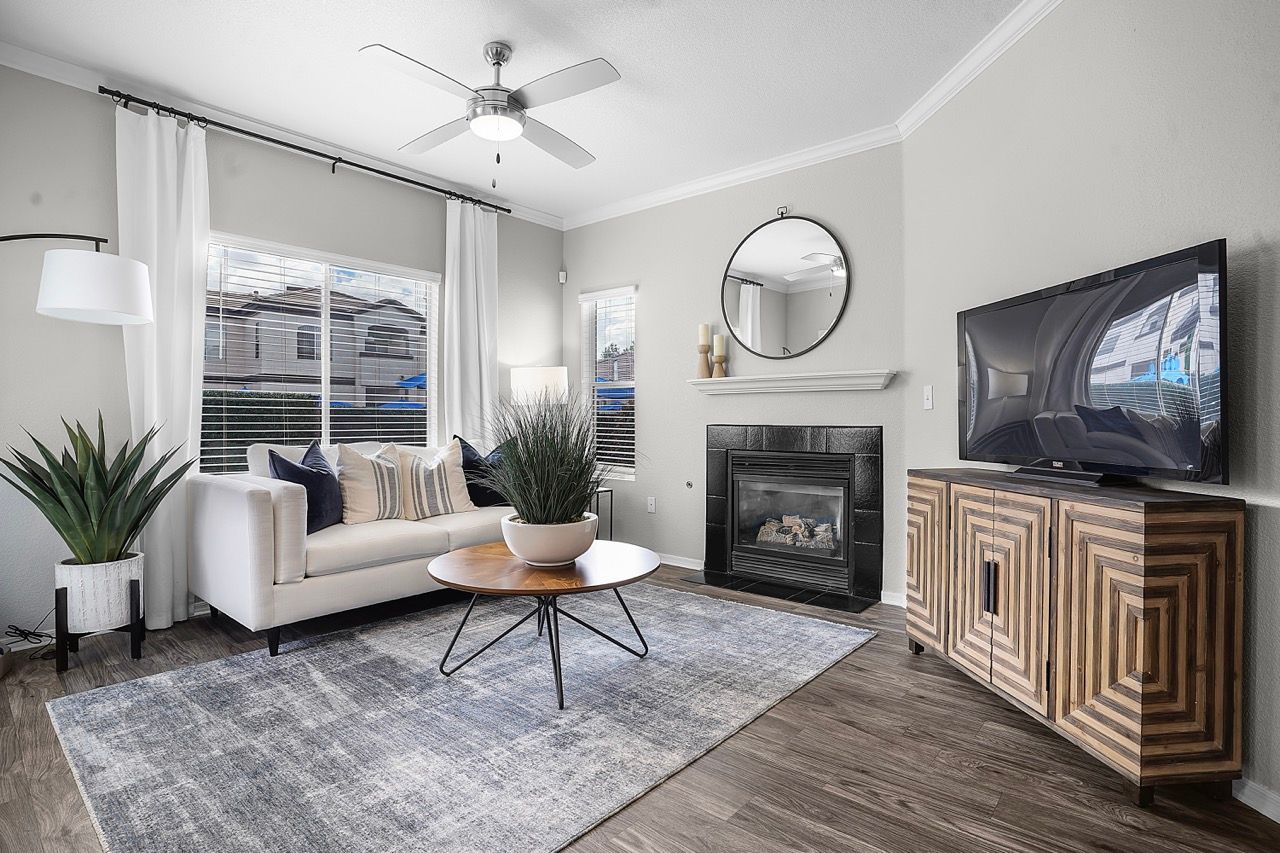 Living room with white sofa, fireplace, round mirror, ceiling fan, and TV on a wooden cabinet at Broadstone Heights in Albuquerque, NM.