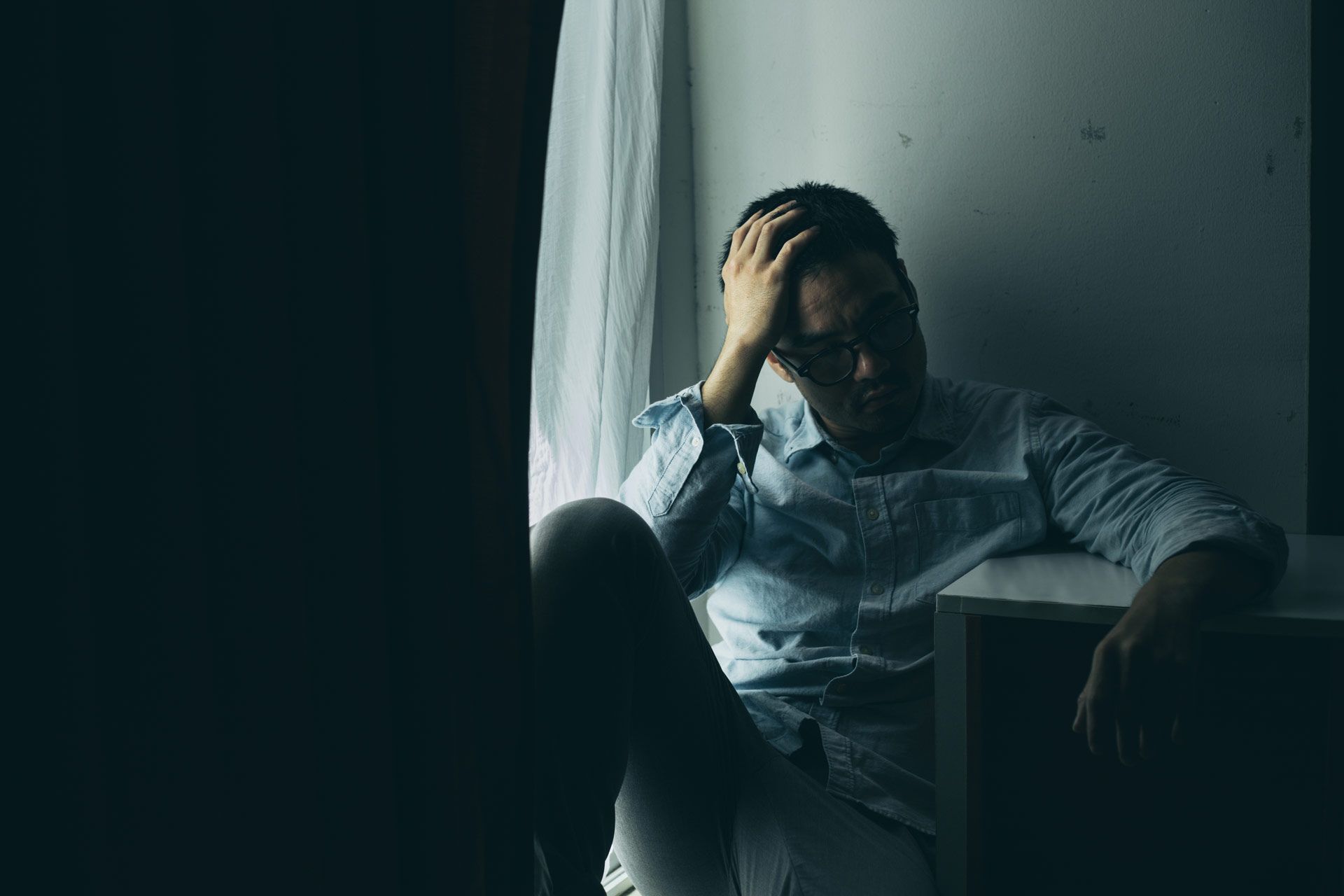 Man sitting by a window, hand on his head, looking stressed in a dim room