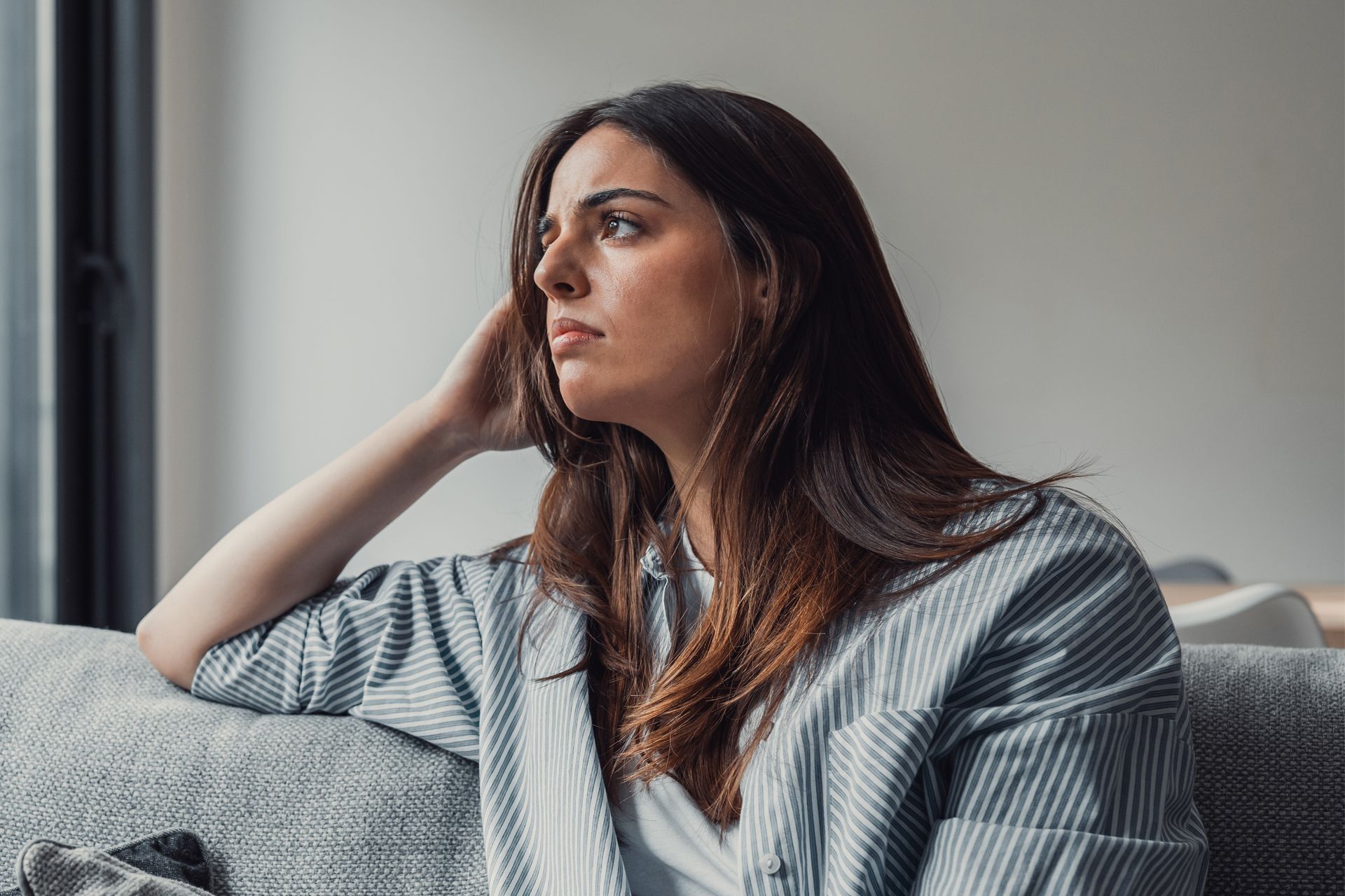 Woman looking out window, resting arm on couch, thoughtful expression.