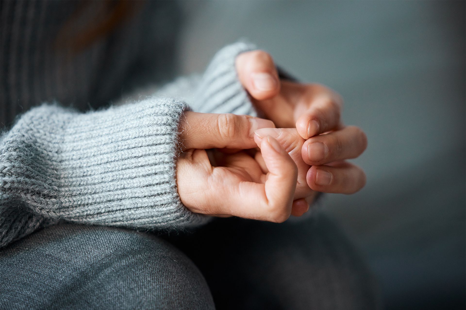 Person's hands clasped together, wearing a gray sweater. Soft focus.