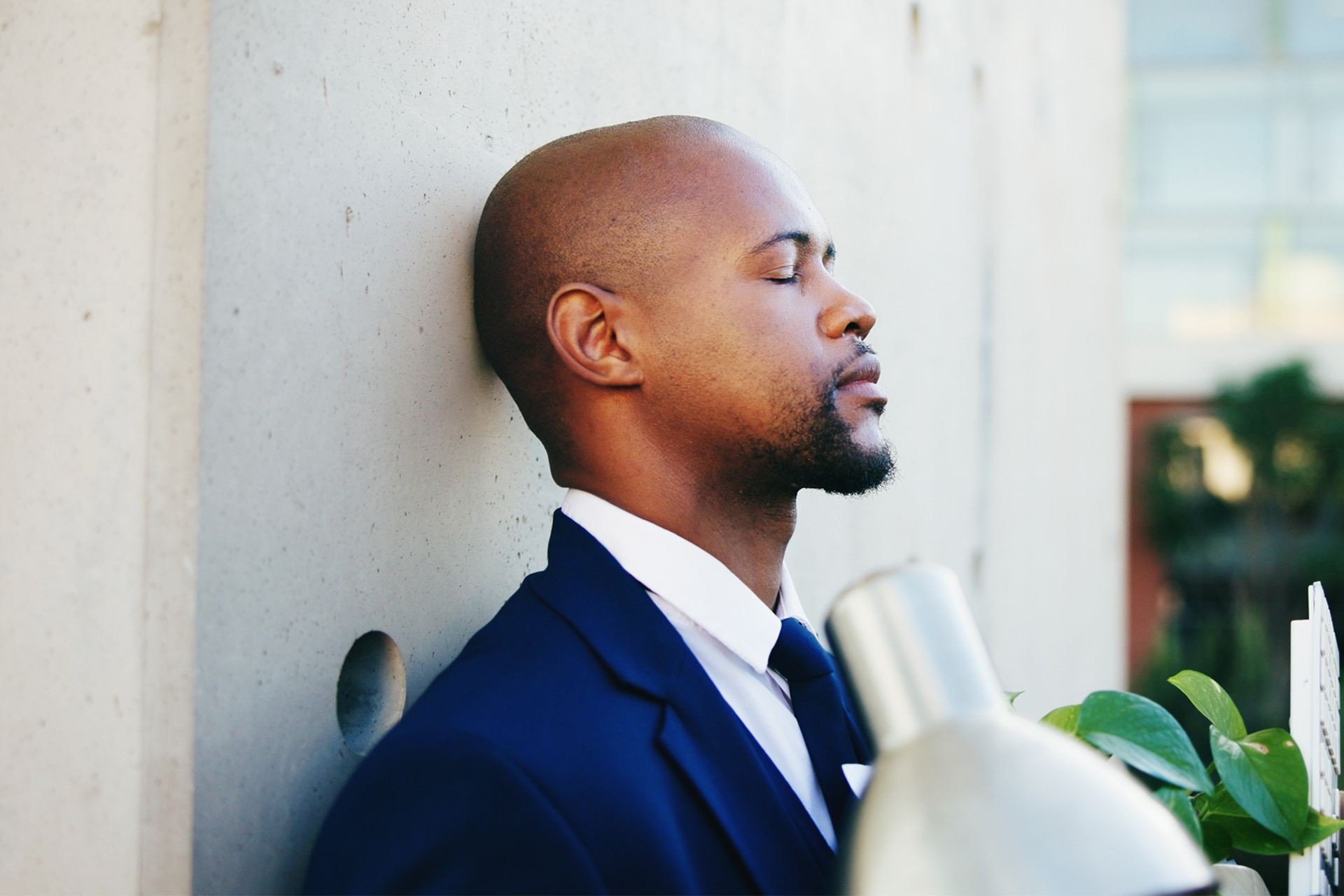 Man with a shaved head in a blue suit leans against a wall, eyes closed, with a contemplative expression.