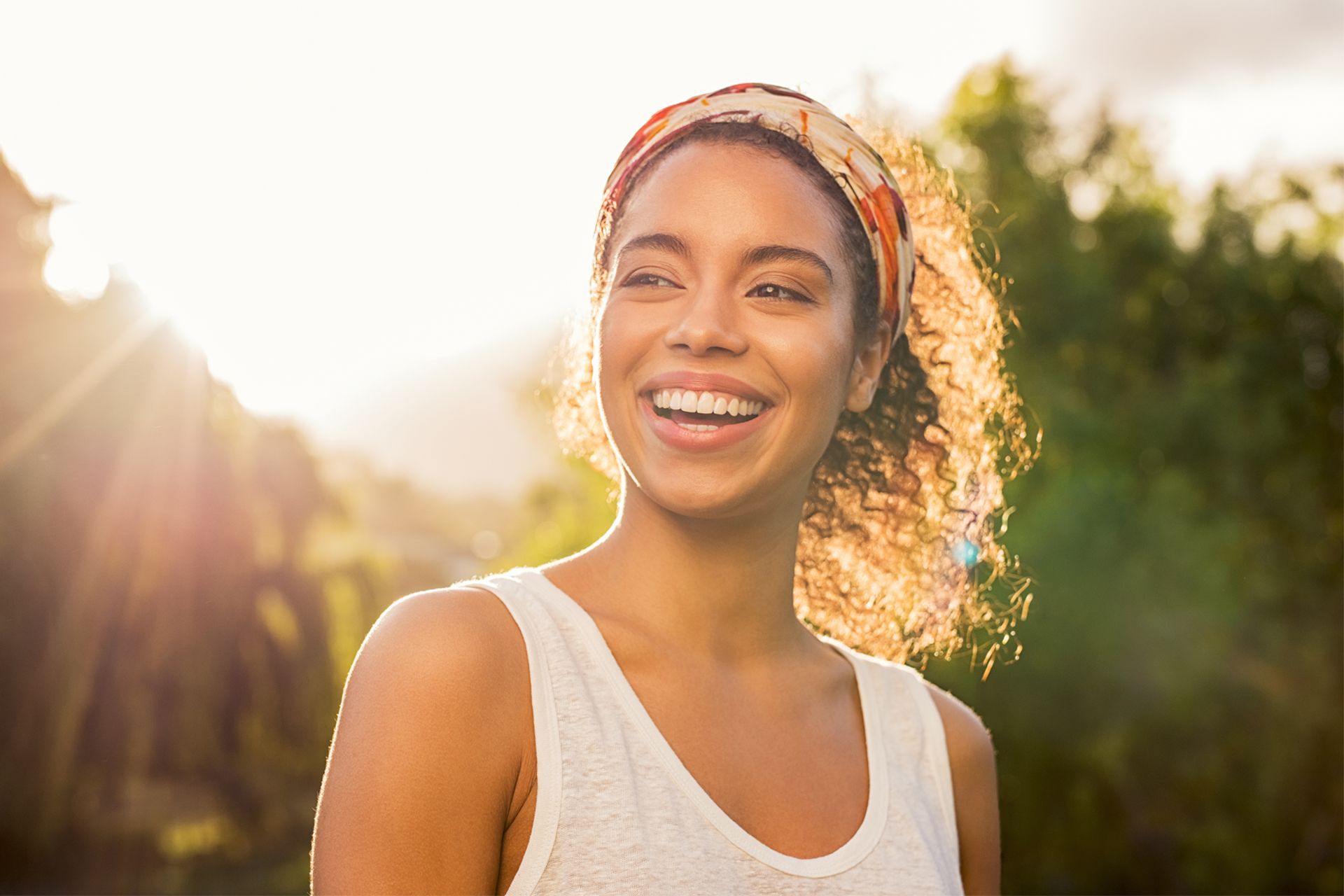 Smiling person outdoors in sunlight, wearing a white tank top and headband.