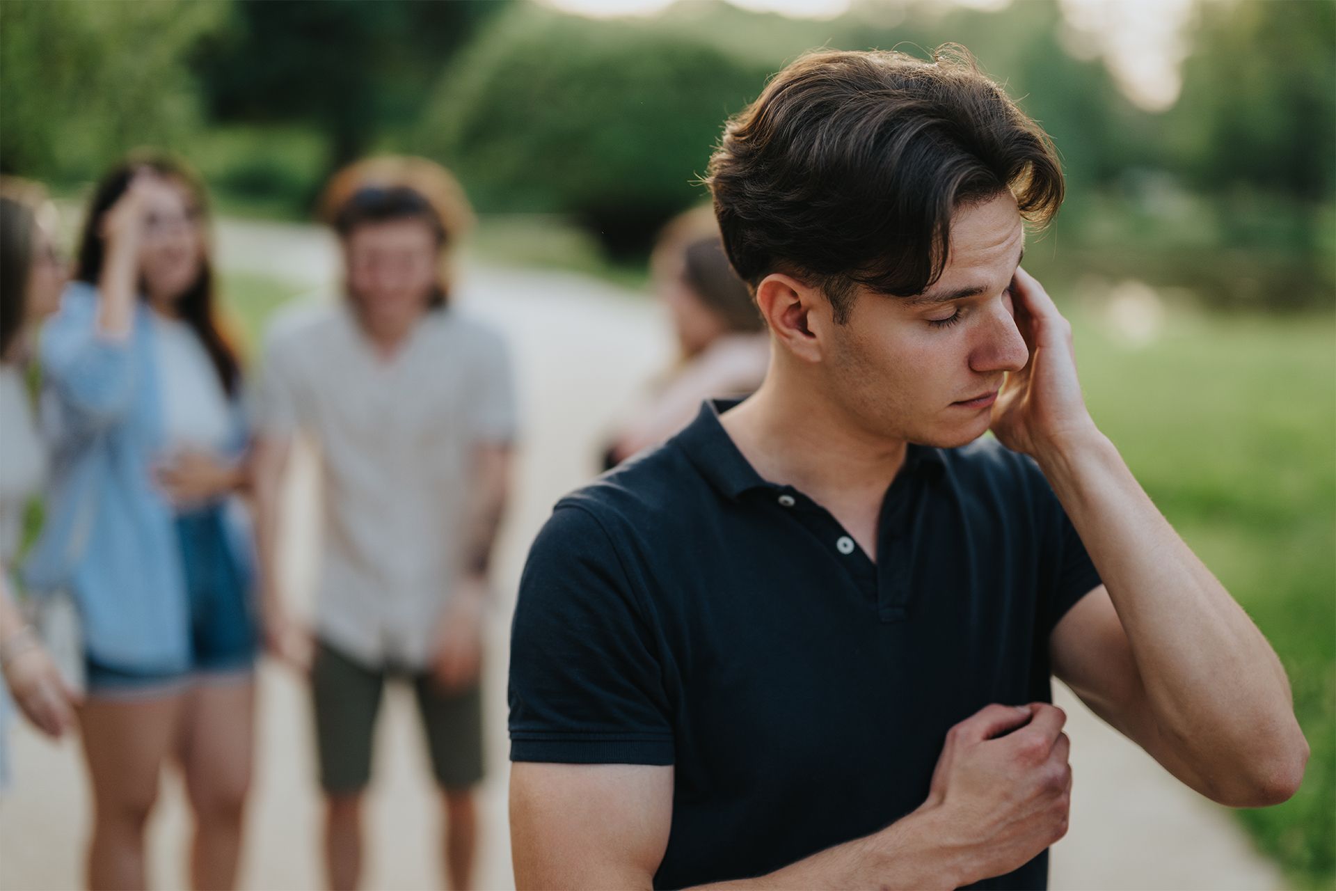 Young man standing outdoors, touching his face while two people walk in the blurred background. Young man standing outdoors, touching his face while two people walk in the blurred background.