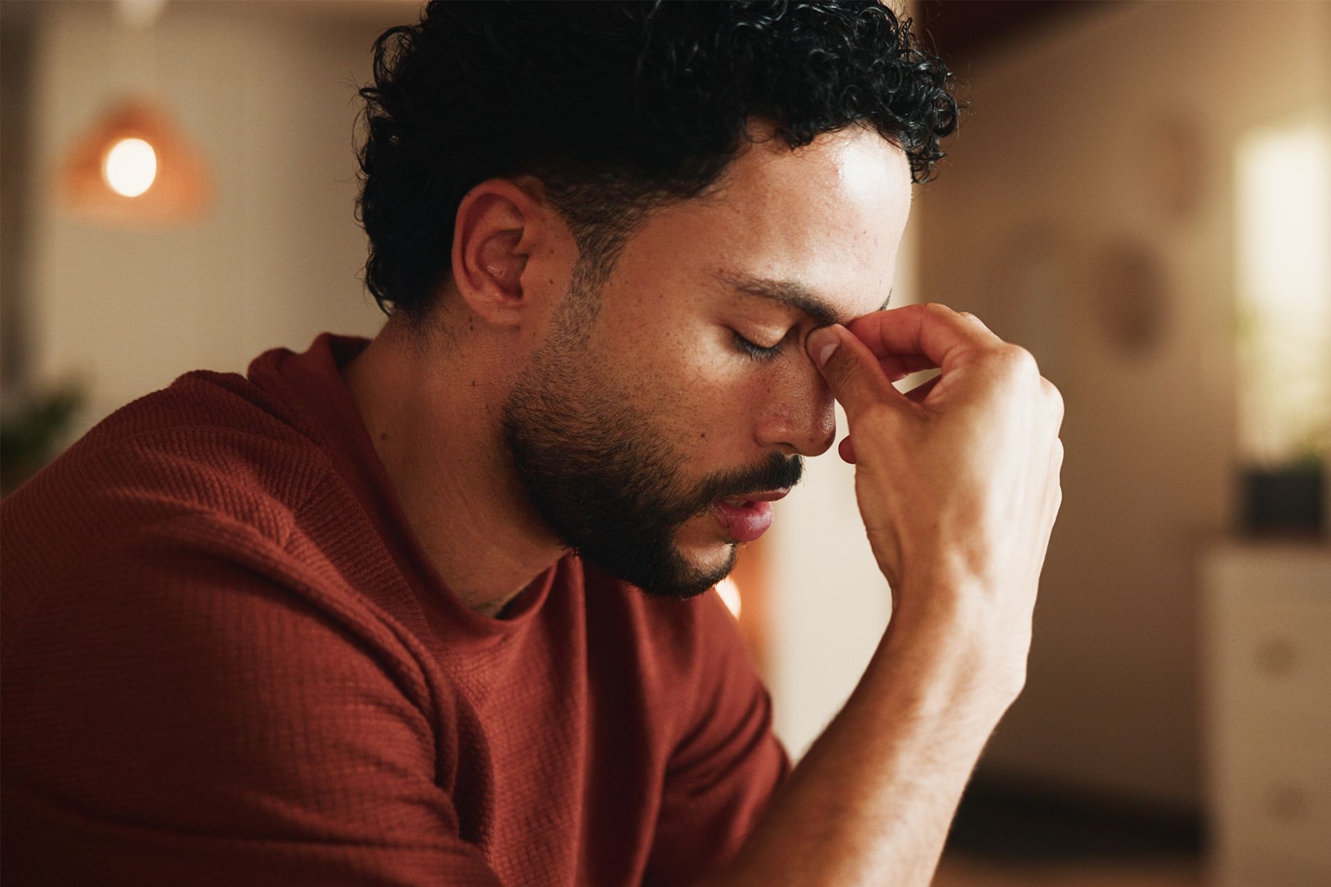 Man sitting indoors with eyes closed, rubbing his forehead thoughtfully.